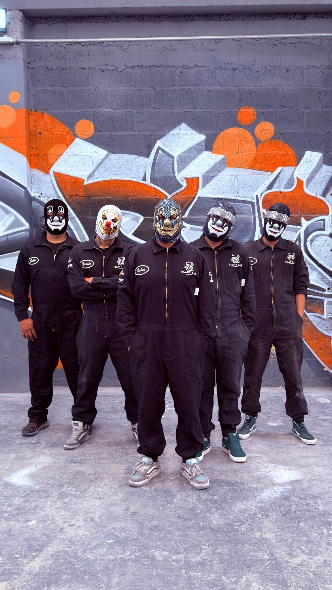 Cinco integrantes de Black Cat Garage con sus máscaras y overoles frente al mural del taller